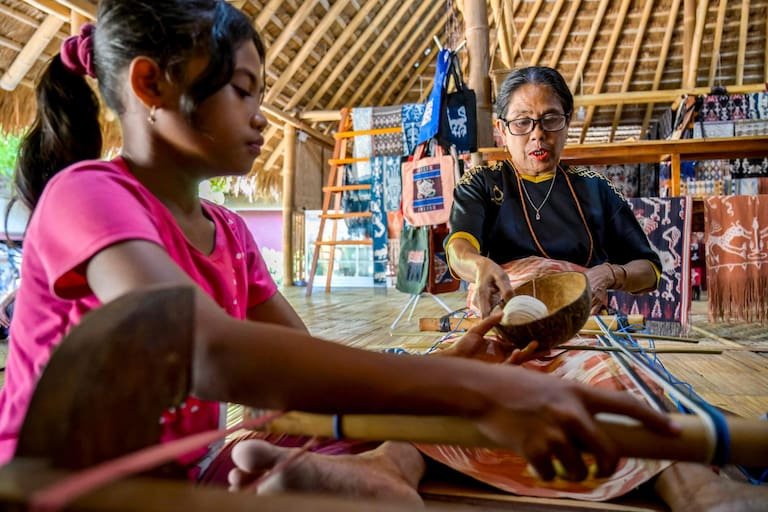 This photo taken on June 24, 2026 shows a girl helping a woman weave traditional ikat fabric, a skill passed down among members of the 'ata' (slaves/servants), at Kampung Raja Prailiu at Waingapu in East Sumba, East Nusa Tenggara. Two hours from the tourist beaches of Bali, slavery is alive and well on the Indonesian island of Sumba, where people are still being born into bondage. Eight of out 10 of Sumba's population of 685,000 people are Christian but its older "marapu" belief system, that blends animism and ancestor worship, has deep roots. It divides society into three castes, "the nobles, called 'maramba' -- often large landowners -- ordinary people called 'kabihu' and the slaves/servants, the atas," said rights activist Martha Hebi, author of several studies on the subject. (Photo by BAY ISMOYO / AFP) / To go with Indonesia-Sumba-slavery, SPECIAL REPORT by Eric Bernaudeau